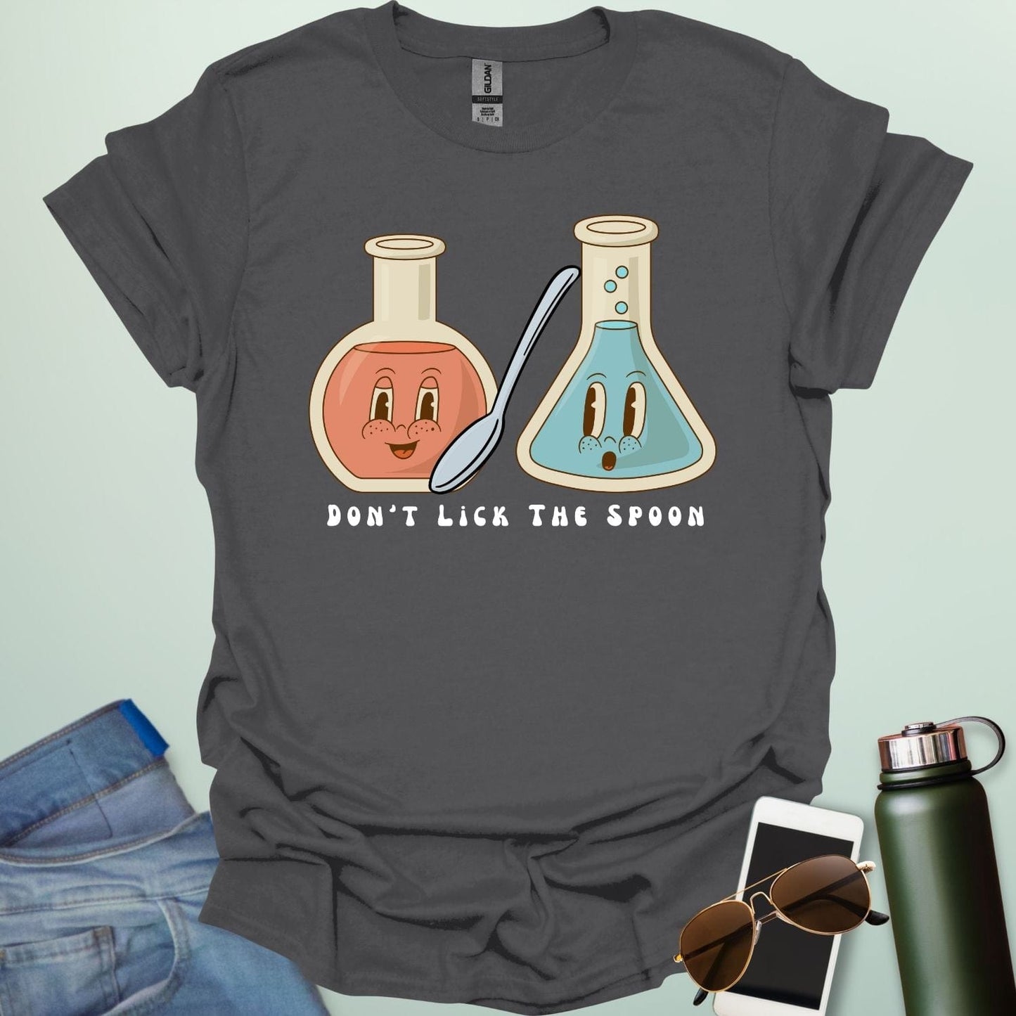 A charcoal grey t-shirt with a cartoon illustration of two lab flasks and a spoon, with the text 'Don't Lick The Spoon.