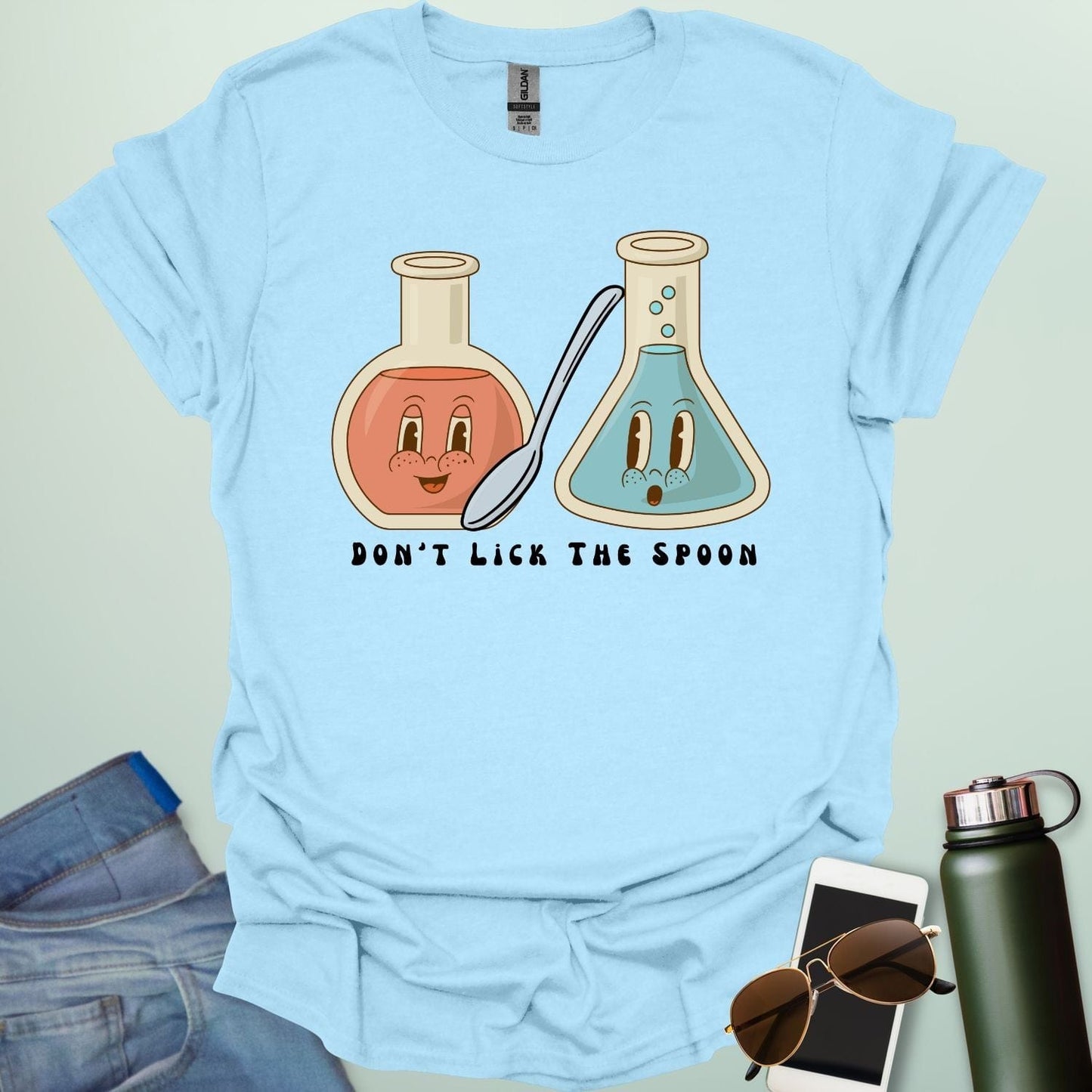A light blue t-shirt with a cartoon illustration of two lab flasks and a spoon, with the text 'Don't Lick The Spoon.