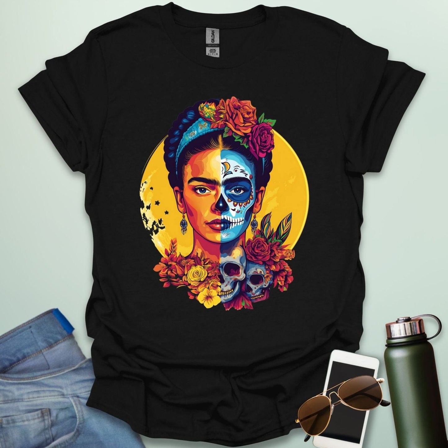 A black t-shirt with a portrait of Frida Kahlo with half of her face as a Dia de los Muertos skull, surrounded by flowers and skulls.