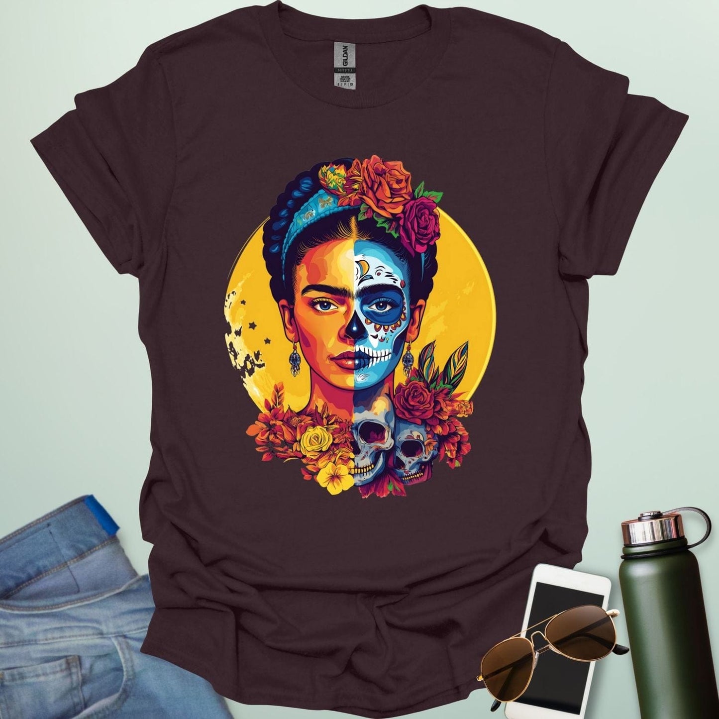 A dark chocolate t-shirt with a portrait of Frida Kahlo with half of her face as a Dia de los Muertos skull, surrounded by flowers and skulls.