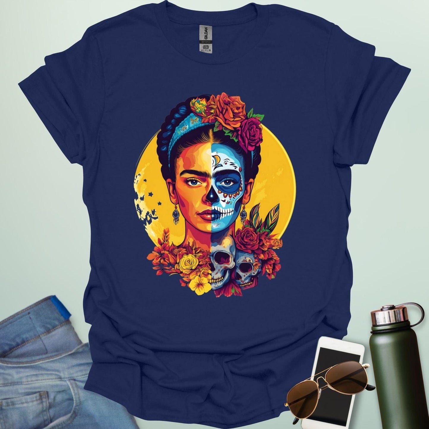A navy blue t-shirt with a portrait of Frida Kahlo with half of her face as a Dia de los Muertos skull, surrounded by flowers and skulls.