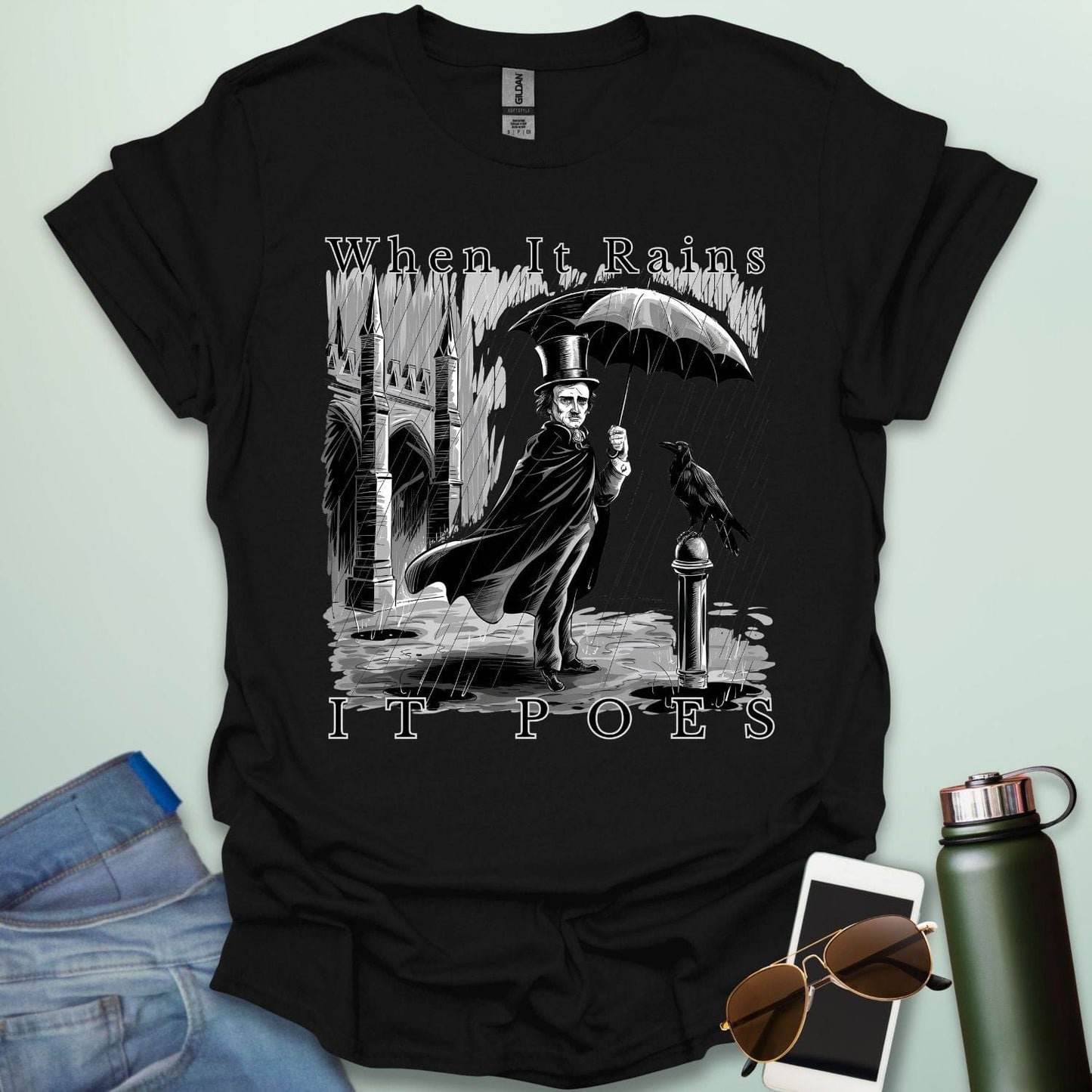 A literary pun t-shirt with a vintage illustration of Edgar Allan Poe holding an umbrella in the rain, next to a raven. The text reads 'When It Rains It Poes.'
