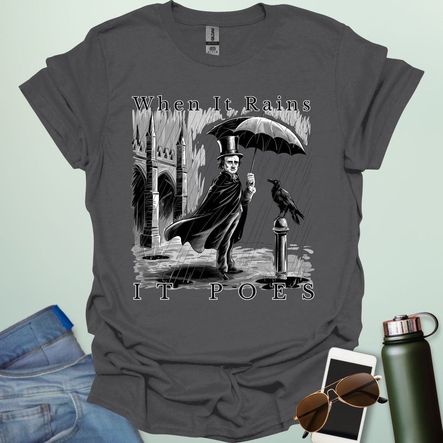 A literary pun t-shirt with a vintage illustration of Edgar Allan Poe holding an umbrella in the rain, next to a raven. The text reads 'When It Rains It Poes.'