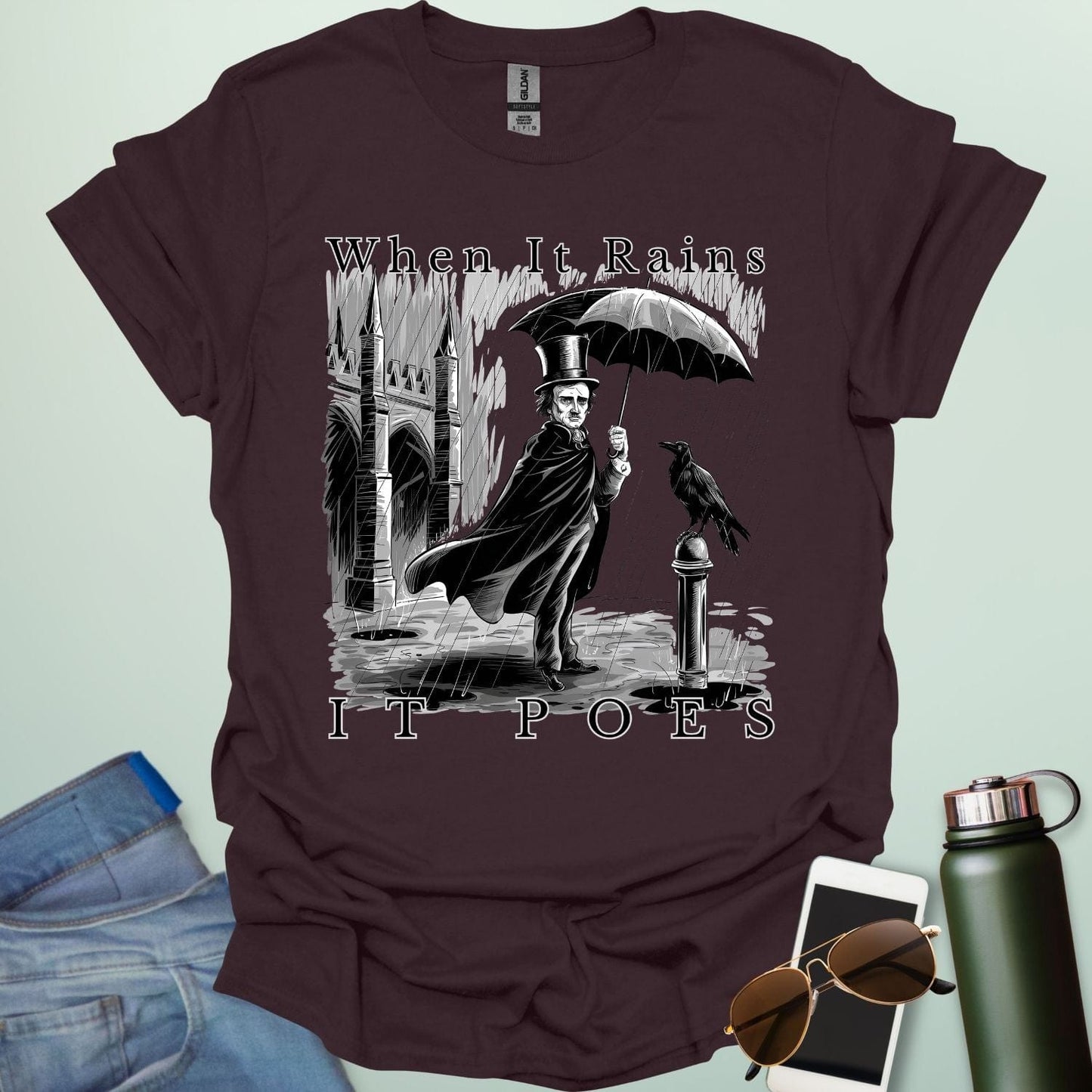 A literary pun t-shirt with a vintage illustration of Edgar Allan Poe holding an umbrella in the rain, next to a raven. The text reads 'When It Rains It Poes.'