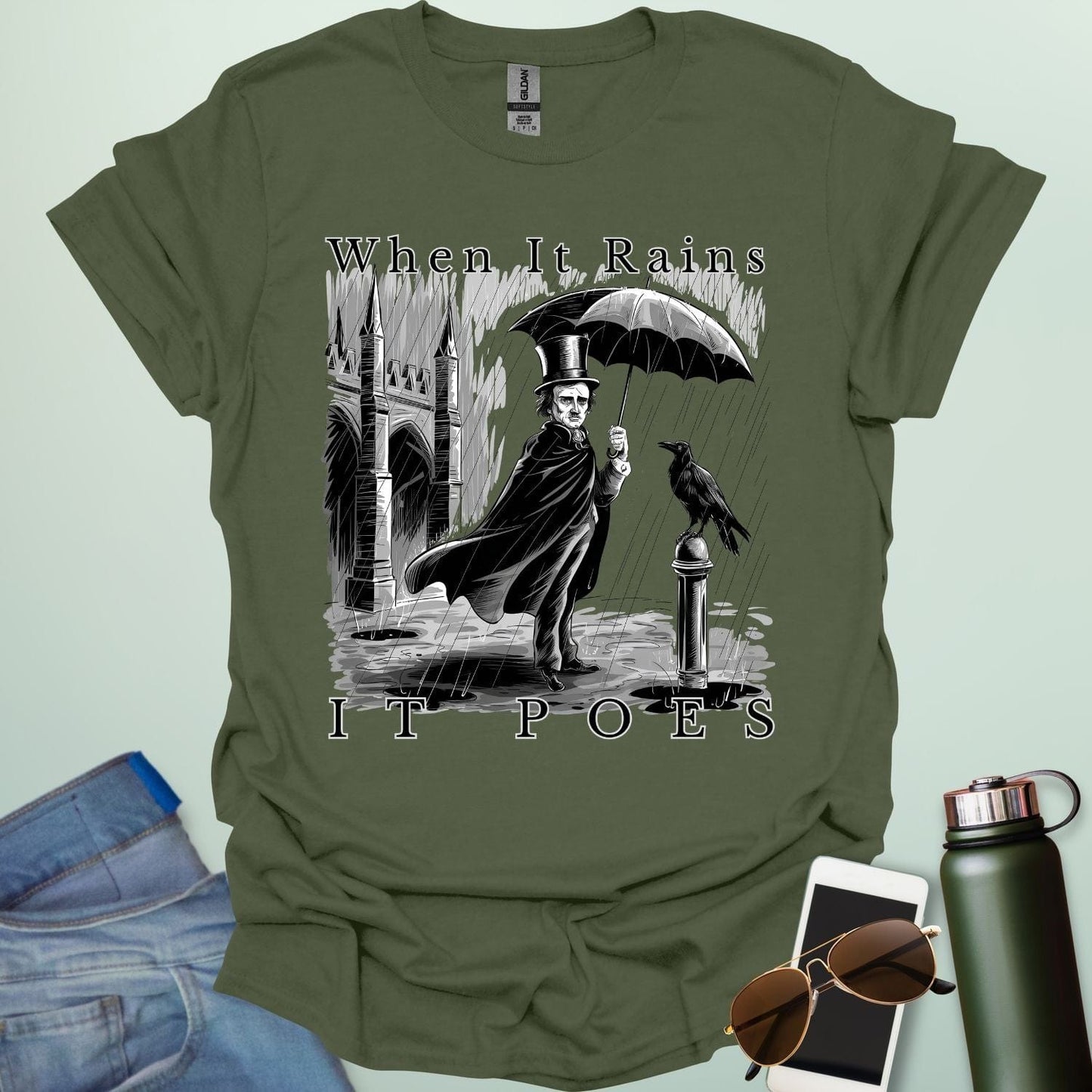 A literary pun t-shirt with a vintage illustration of Edgar Allan Poe holding an umbrella in the rain, next to a raven. The text reads 'When It Rains It Poes.'