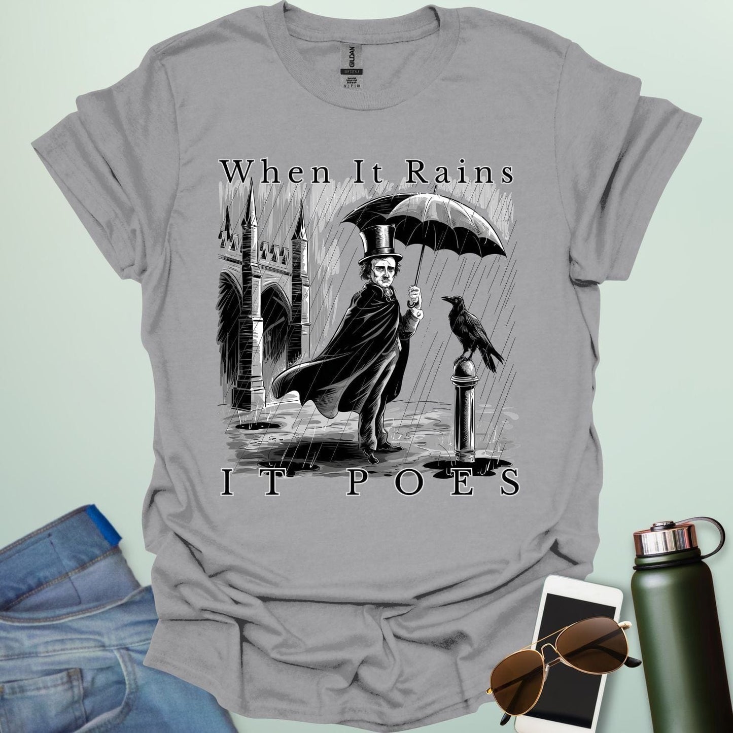 A literary pun t-shirt with a vintage illustration of Edgar Allan Poe holding an umbrella in the rain, next to a raven. The text reads 'When It Rains It Poes.'