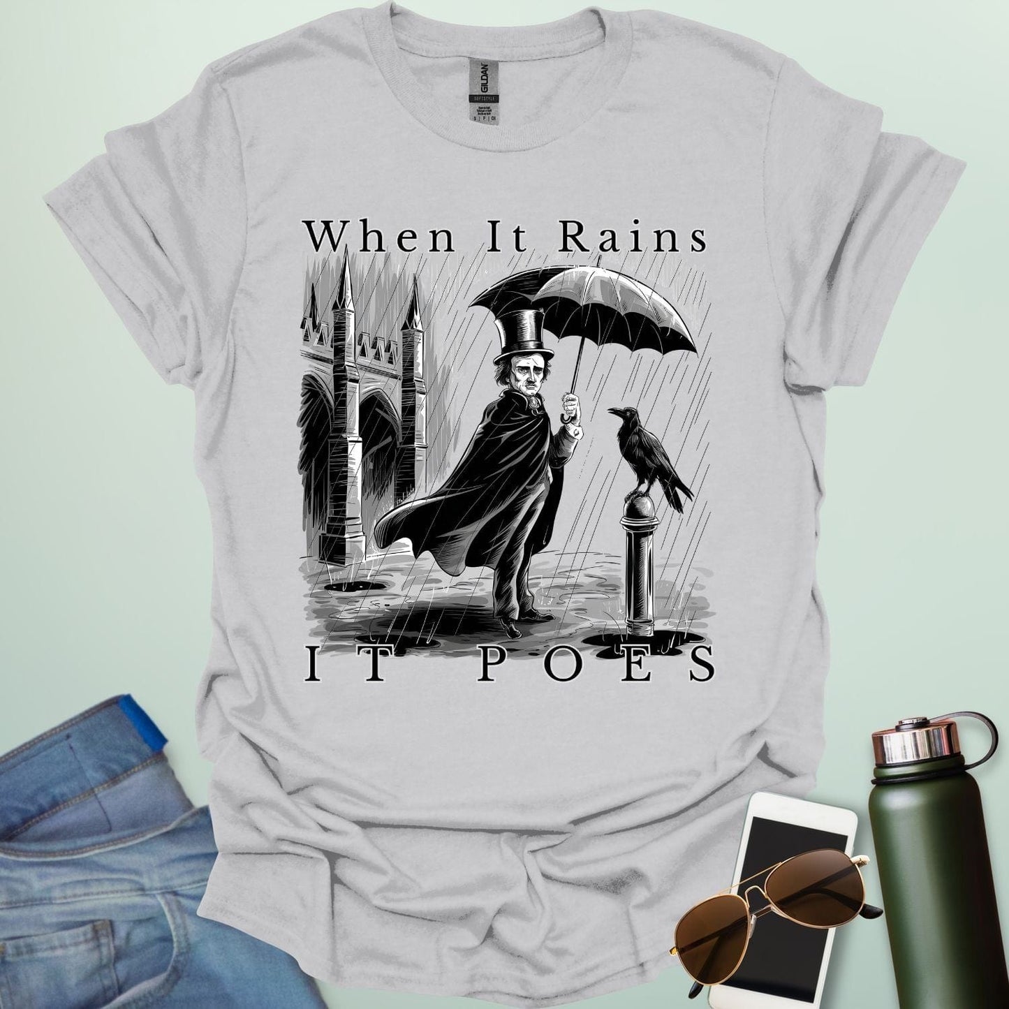 A literary pun t-shirt with a vintage illustration of Edgar Allan Poe holding an umbrella in the rain, next to a raven. The text reads 'When It Rains It Poes.'