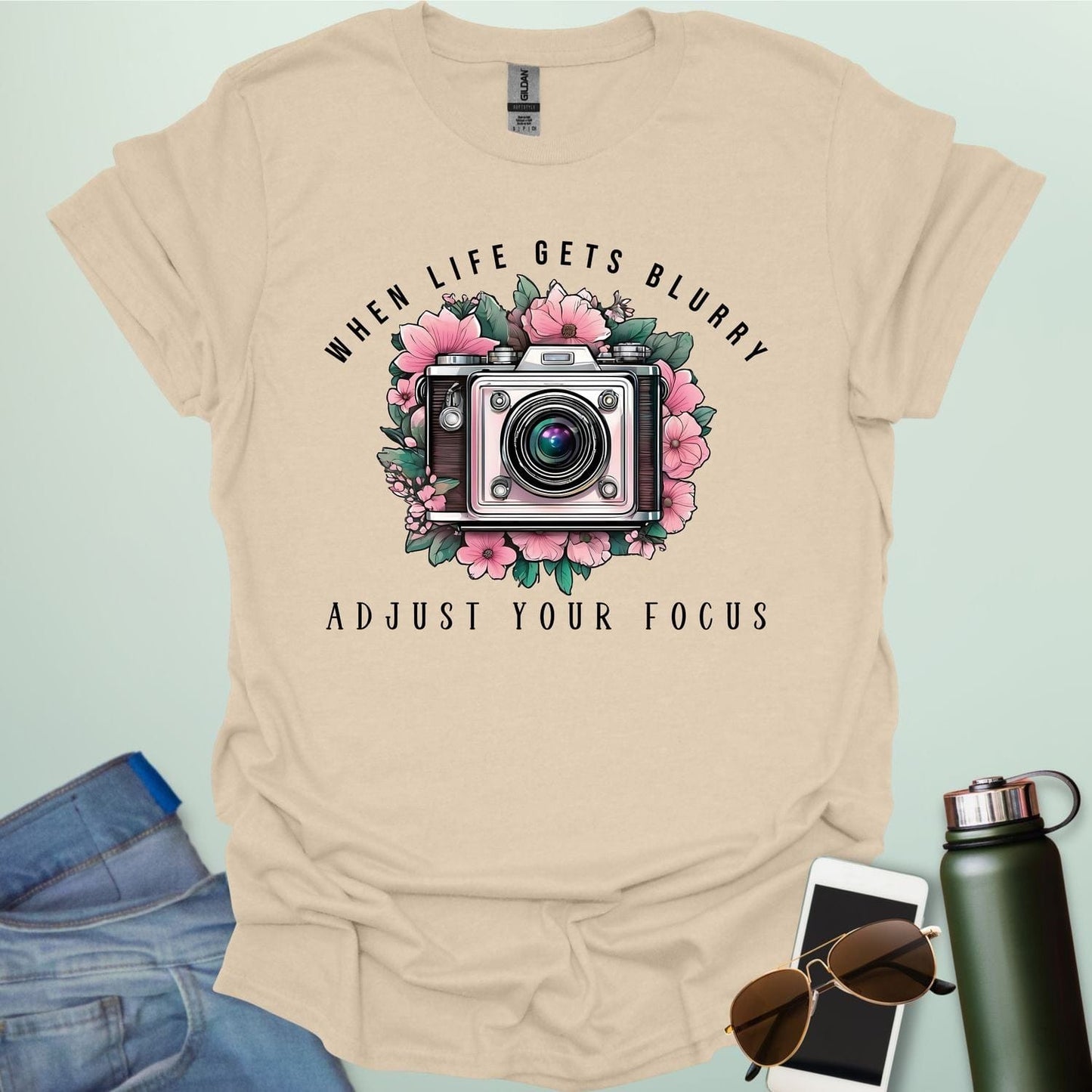 A vintage camera surrounded by pink flowers and green leaves, with the text 'WHEN LIFE GETS BLURRY' arched above and 'ADJUST YOUR FOCUS' below.