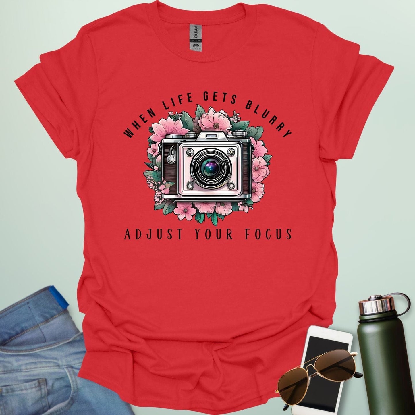 A vintage camera surrounded by pink flowers and green leaves, with the text 'WHEN LIFE GETS BLURRY' arched above and 'ADJUST YOUR FOCUS' below.