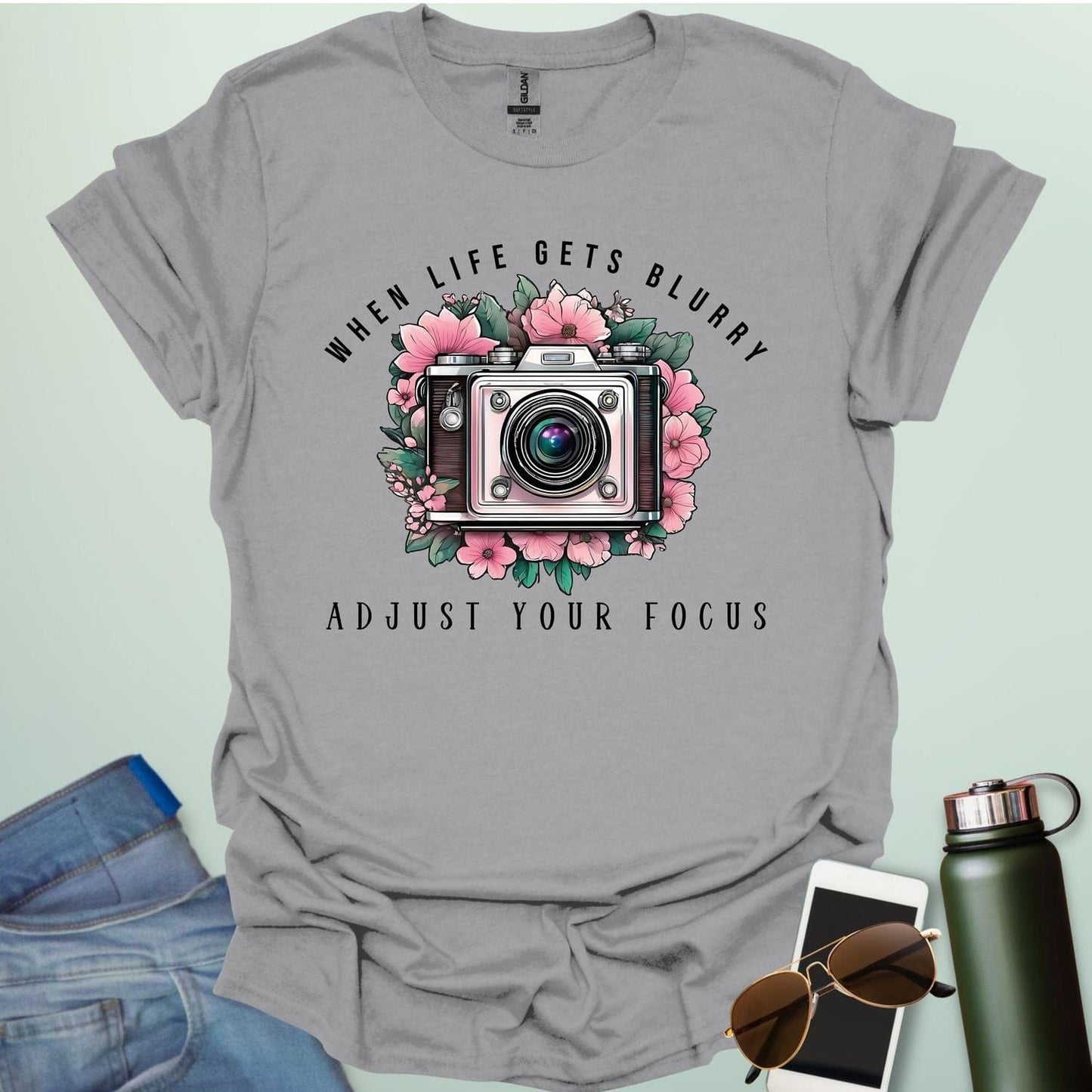 A vintage camera surrounded by pink flowers and green leaves, with the text 'WHEN LIFE GETS BLURRY' arched above and 'ADJUST YOUR FOCUS' below.