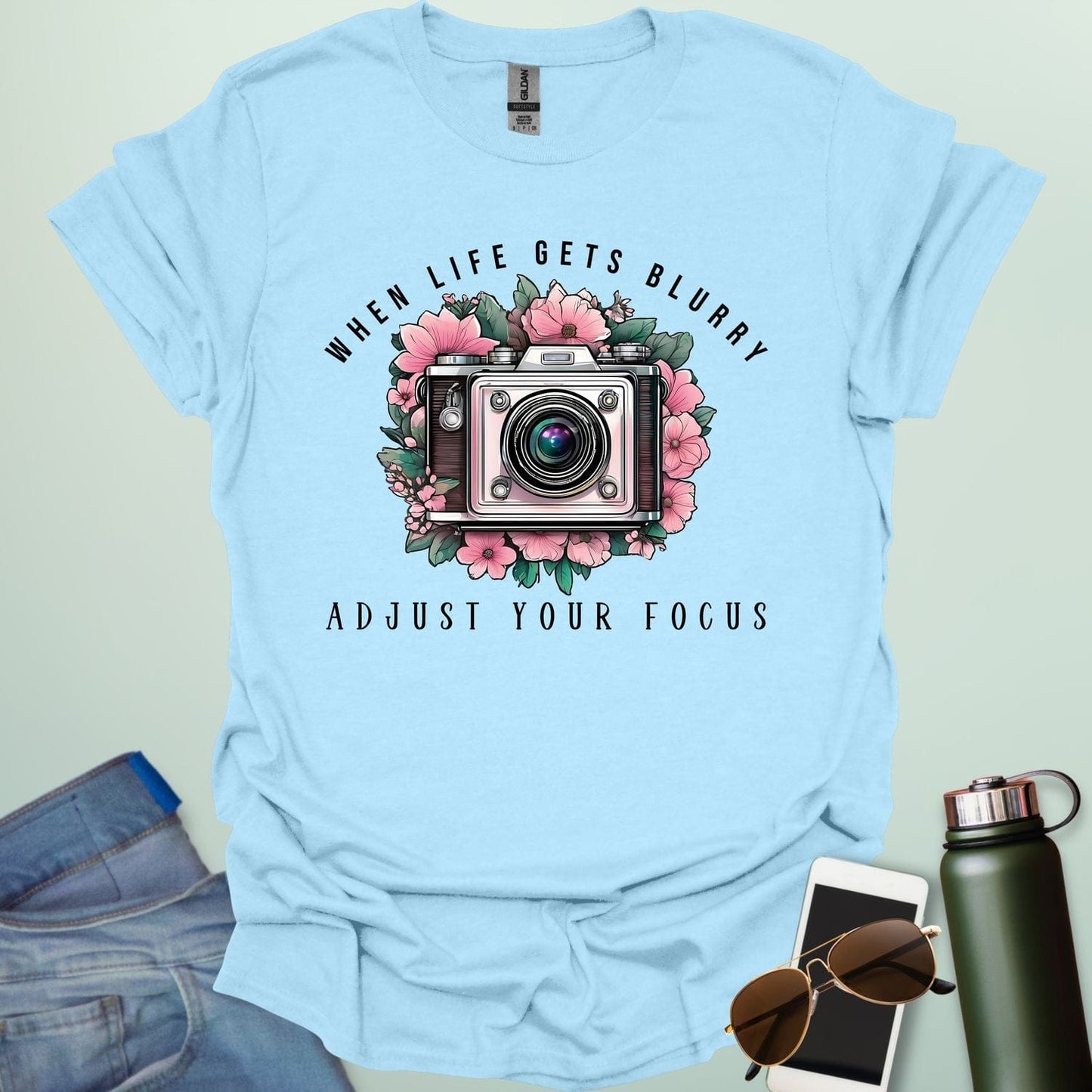 A vintage camera surrounded by pink flowers and green leaves, with the text 'WHEN LIFE GETS BLURRY' arched above and 'ADJUST YOUR FOCUS' below.