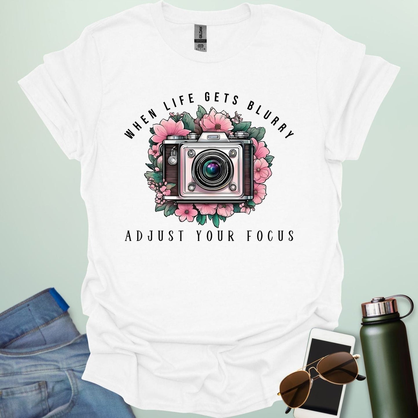 A vintage camera surrounded by pink flowers and green leaves, with the text 'WHEN LIFE GETS BLURRY' arched above and 'ADJUST YOUR FOCUS' below.