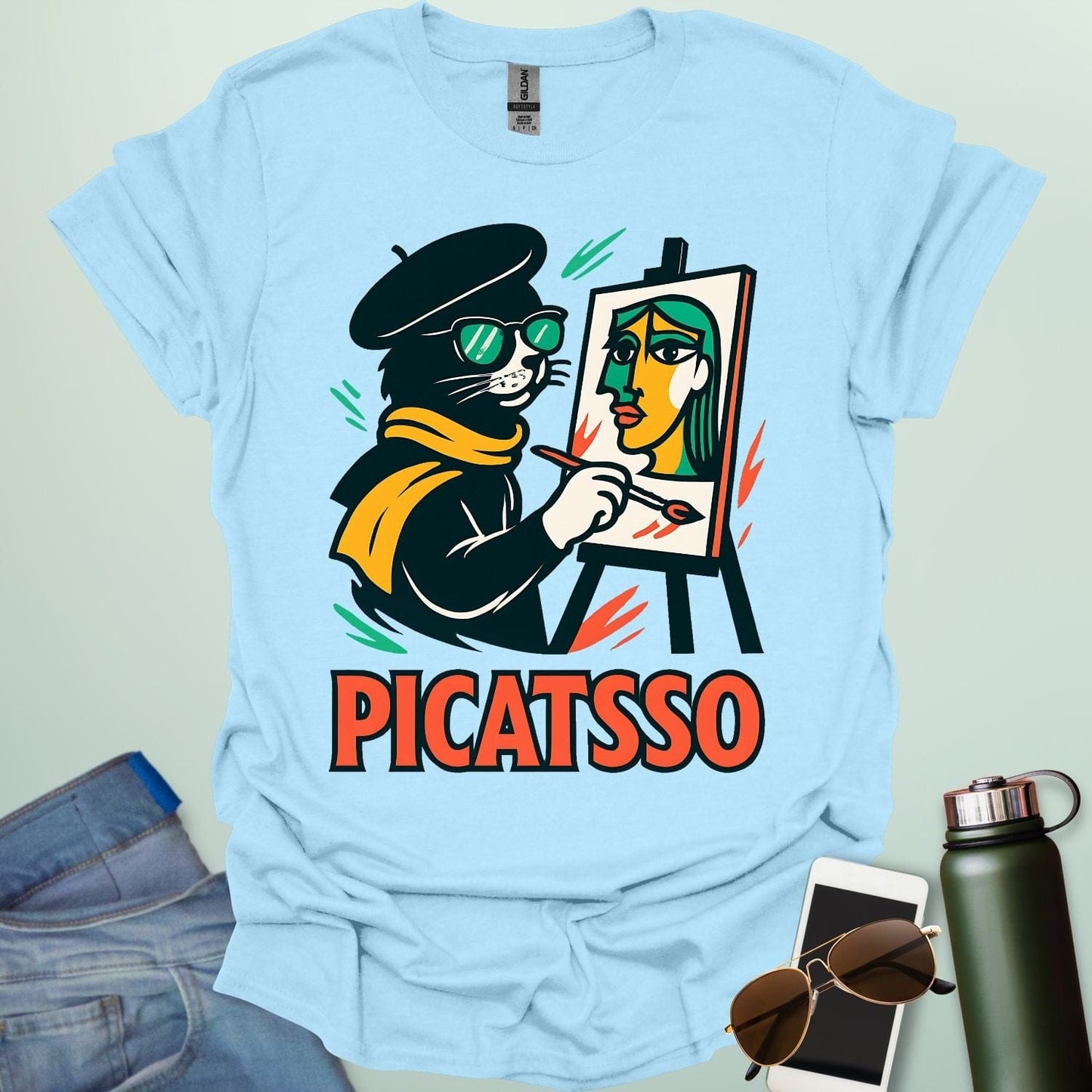 A light blue t-shirt with a cartoon of a cat painting a Picasso-style portrait on an easel and the text 'PICATSSO' below.