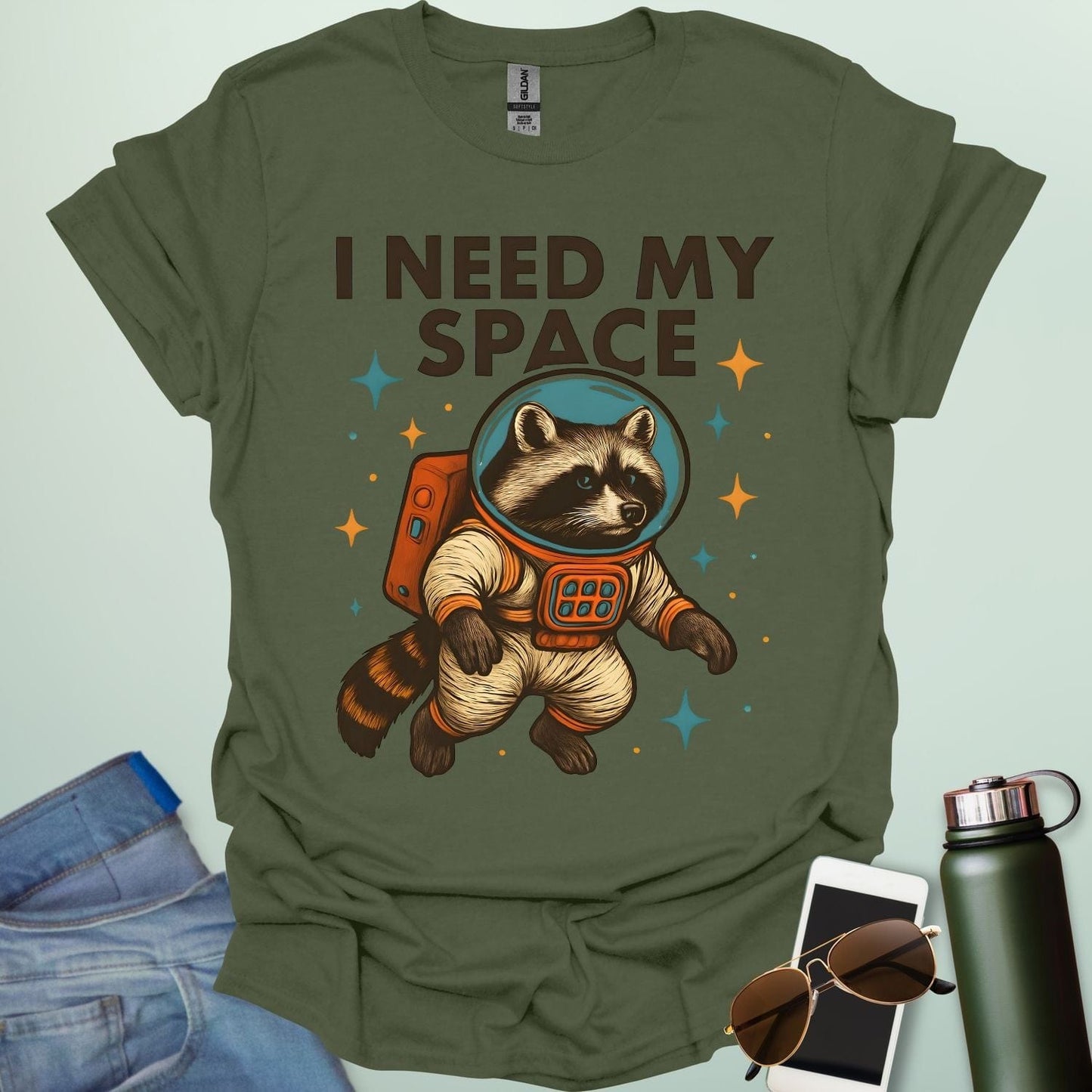 A military green t-shirt with a cartoon illustration of a raccoon in an astronaut suit and the text 'I NEED MY SPACE.'
