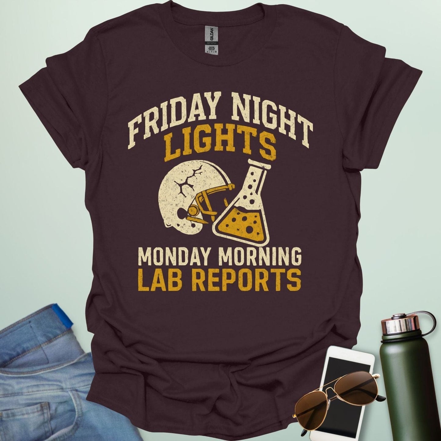 A dark chocolate t-shirt with a vintage football helmet and a cracked lab flask, with the text 'FRIDAY NIGHT LIGHTS' and 'MONDAY MORNING LAB REPORTS.'