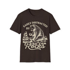 Linocut-style illustration of a bear sitting down examining a stone beside a cairn stack, framed by text reading 'EASILY DISTRACTED BY ROCKS'.