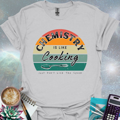 The text 'Chemistry is like Cooking - Just don't lick the spoon' in a distressed, grunge font, with a spoon graphic and a retro-style sunset background.