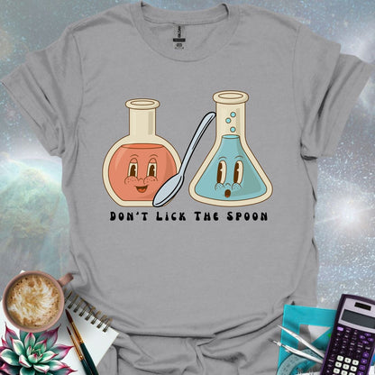 T-shirt design featuring two cartoonish retro beakers, one smiling, one surprised, with a spoon and the text 'Don't Lick The Spoon'.