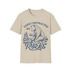 Linocut-style illustration of a bear sitting down examining a stone beside a cairn stack, framed by text reading 'EASILY DISTRACTED BY ROCKS'.