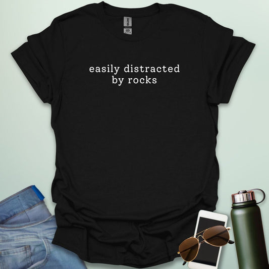 EASYLY DISTRACTED BY ROCKS t-shirt with distressed uppercase typography and small rock icons beside ‘BY’ — minimalist geology humor tee.