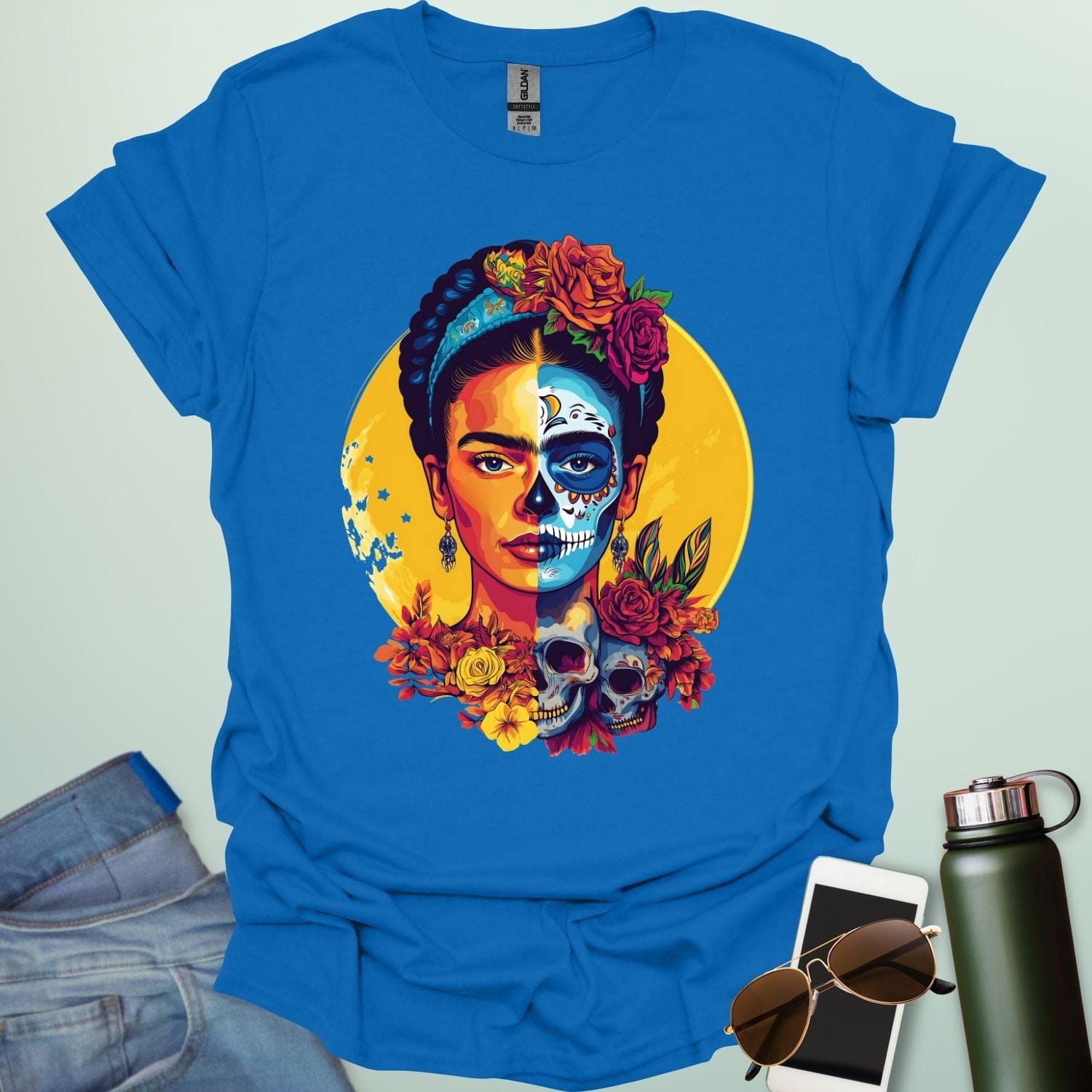 A royal blue t-shirt with a portrait of Frida Kahlo with half of her face as a Dia de los Muertos skull, surrounded by flowers and skulls.