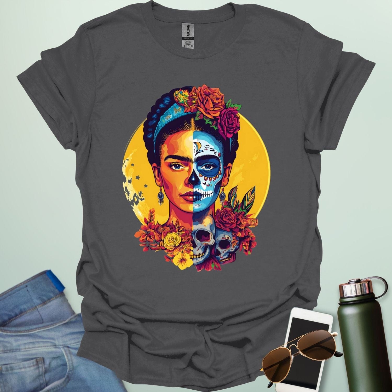 A charcoal grey t-shirt with a portrait of Frida Kahlo with half of her face as a Dia de los Muertos skull, surrounded by flowers and skulls.