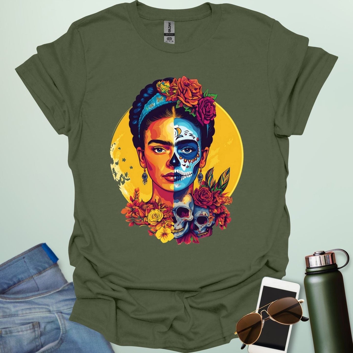 A military green t-shirt with a portrait of Frida Kahlo with half of her face as a Dia de los Muertos skull, surrounded by flowers and skulls.