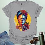 An ice grey  t-shirt with a portrait of Frida Kahlo with half of her face as a Dia de los Muertos skull, surrounded by flowers and skulls.