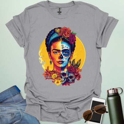 An ice grey  t-shirt with a portrait of Frida Kahlo with half of her face as a Dia de los Muertos skull, surrounded by flowers and skulls.