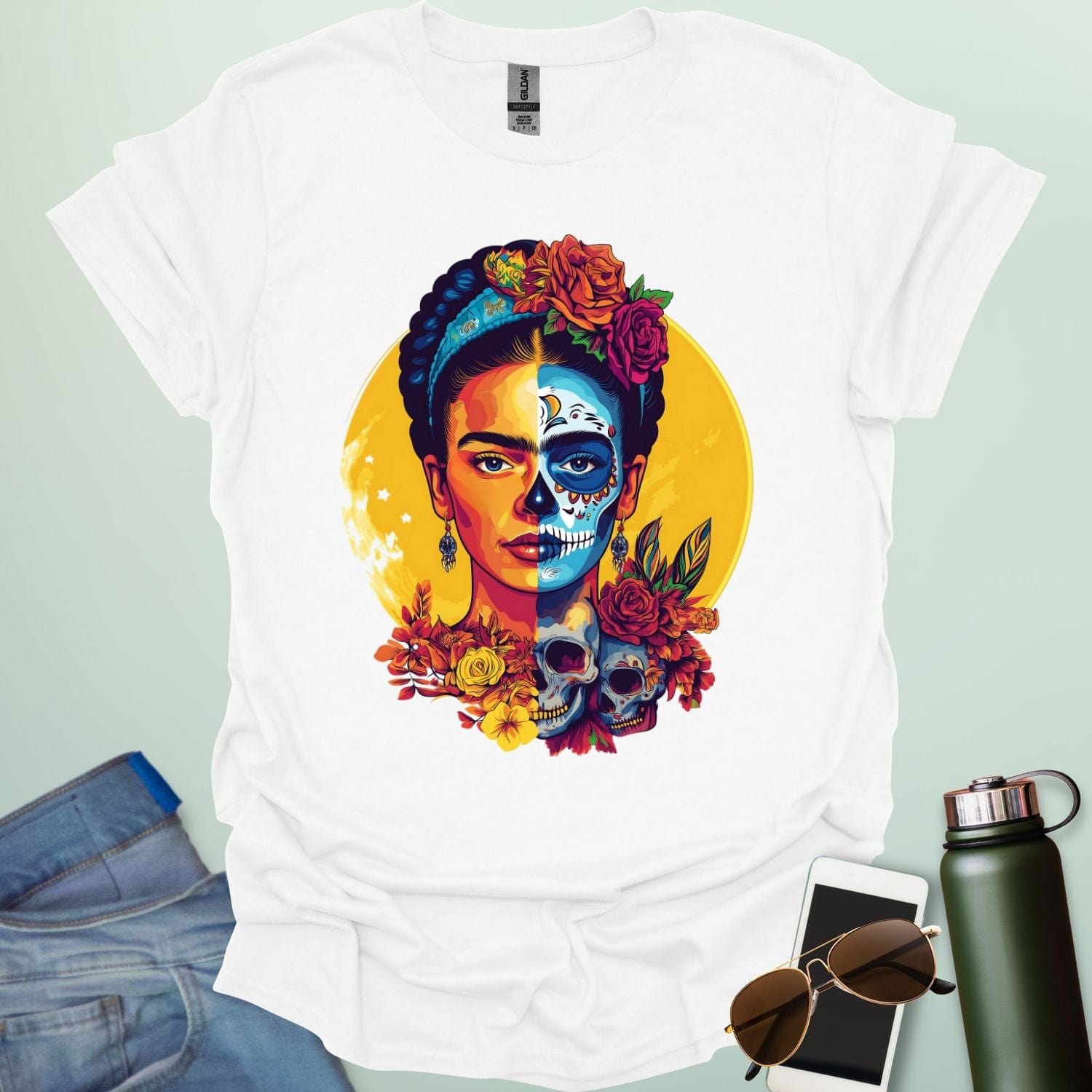 A white t-shirt with a portrait of Frida Kahlo with half of her face as a Dia de los Muertos skull, surrounded by flowers and skulls.