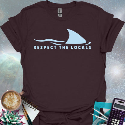 A blue shark fin rising above blue ocean waves, with the text 'RESPECT THE LOCALS' below.