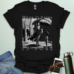 A literary pun t-shirt with a vintage illustration of Edgar Allan Poe holding an umbrella in the rain, next to a raven. The text reads 'When It Rains It Poes.'