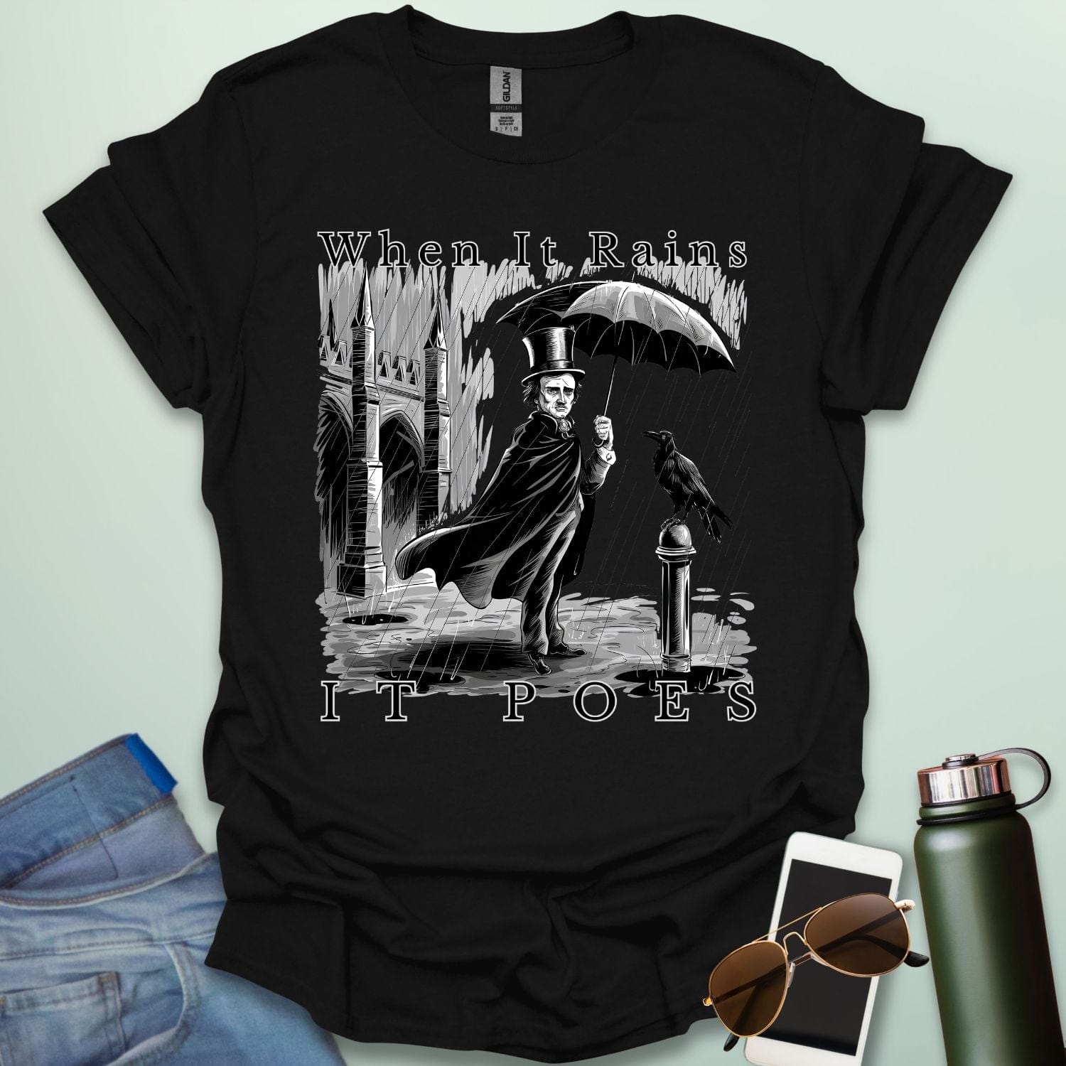 A literary pun t-shirt with a vintage illustration of Edgar Allan Poe holding an umbrella in the rain, next to a raven. The text reads 'When It Rains It Poes.'