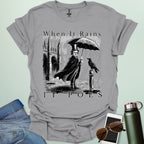 A literary pun t-shirt with a vintage illustration of Edgar Allan Poe holding an umbrella in the rain, next to a raven. The text reads 'When It Rains It Poes.'