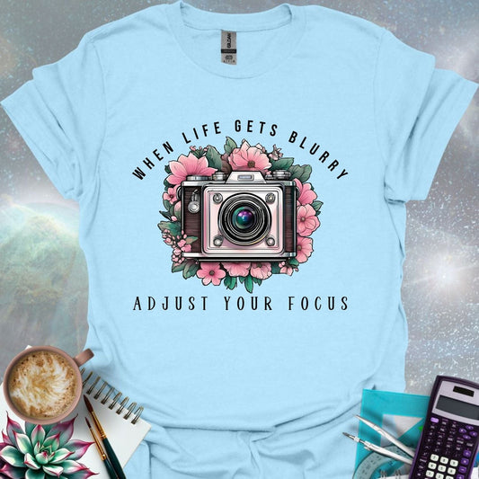 A vintage camera surrounded by pink flowers and green leaves, with the text 'WHEN LIFE GETS BLURRY' arched above and 'ADJUST YOUR FOCUS' below.