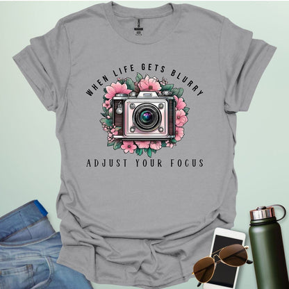A vintage camera surrounded by pink flowers and green leaves, with the text 'WHEN LIFE GETS BLURRY' arched above and 'ADJUST YOUR FOCUS' below.