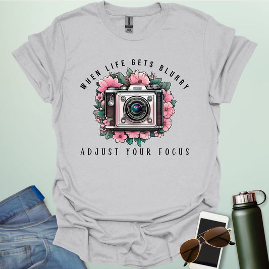 A vintage camera surrounded by pink flowers and green leaves, with the text 'WHEN LIFE GETS BLURRY' arched above and 'ADJUST YOUR FOCUS' below.