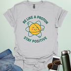 A cheerful graphic design featuring a smiling yellow proton character with orbiting electrons and the text "BE LIKE A PROTON STAY POSITIVE."