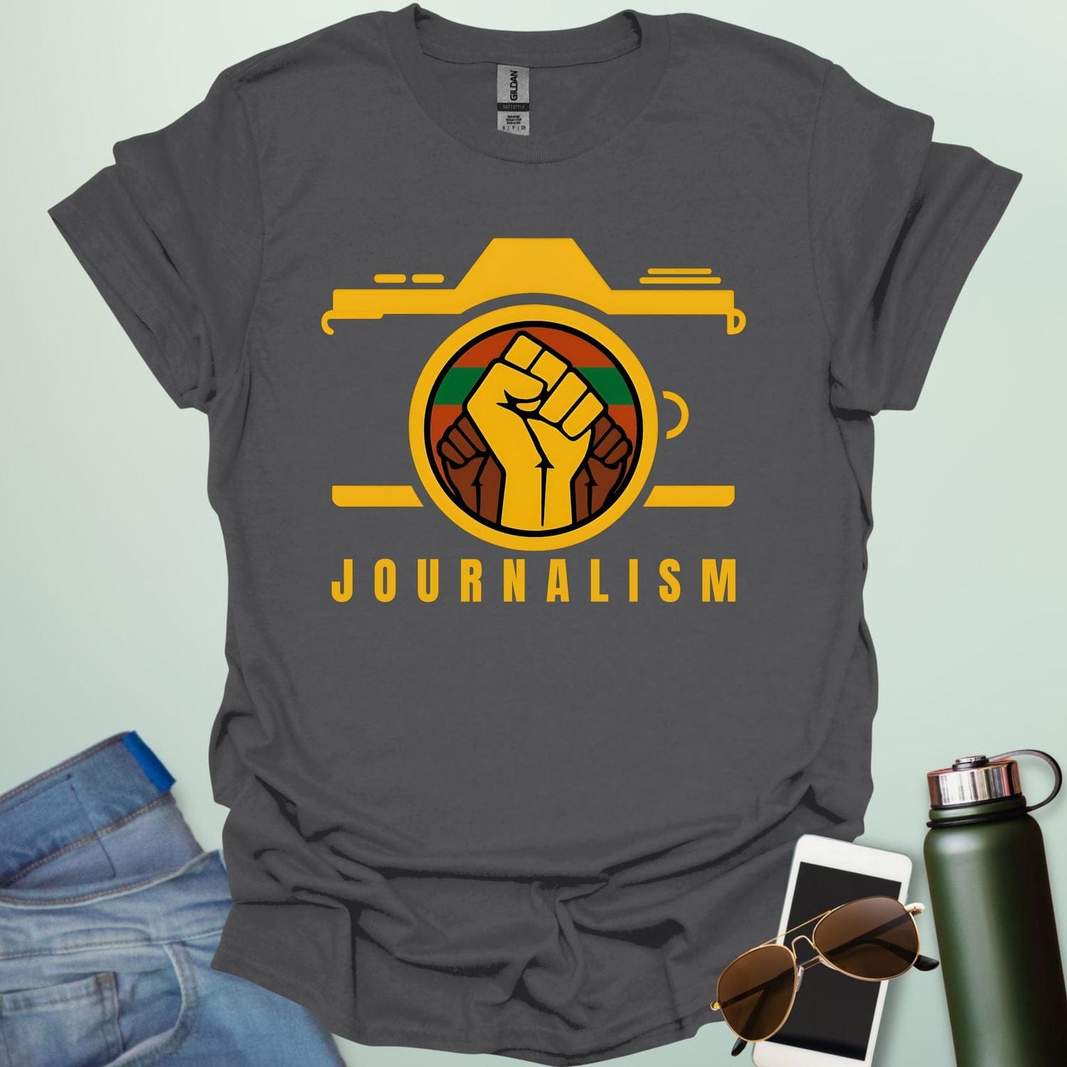 A graphic t-shirt design showing a stylized camera with a powerful raised fist inside its lens. The fist has a green, red, and black striped background. Below the image, the word "JOURNALISM" is written in yellow capital letters.