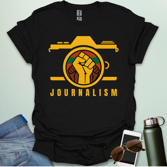 A graphic t-shirt design showing a stylized camera with a powerful raised fist inside its lens. The fist has a green, red, and black striped background. Below the image, the word "JOURNALISM" is written in yellow capital letters.