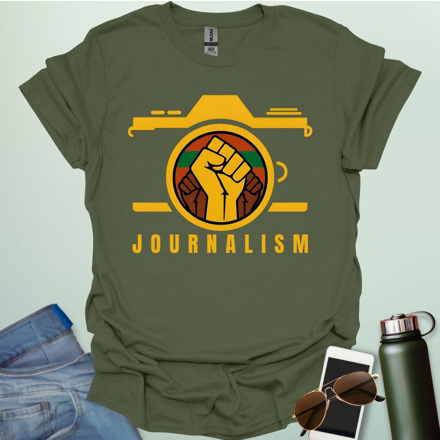 A graphic t-shirt design showing a stylized camera with a powerful raised fist inside its lens. The fist has a green, red, and black striped background. Below the image, the word "JOURNALISM" is written in yellow capital letters.