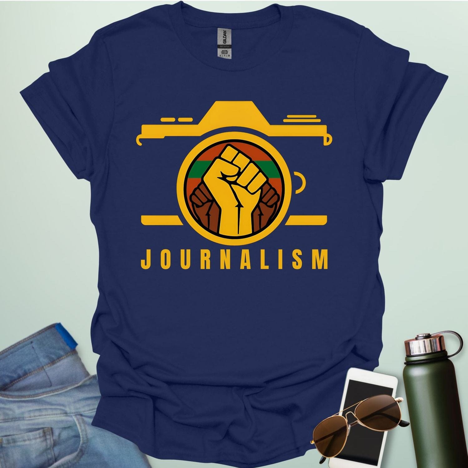 A graphic t-shirt design showing a stylized camera with a powerful raised fist inside its lens. The fist has a green, red, and black striped background. Below the image, the word "JOURNALISM" is written in yellow capital letters.