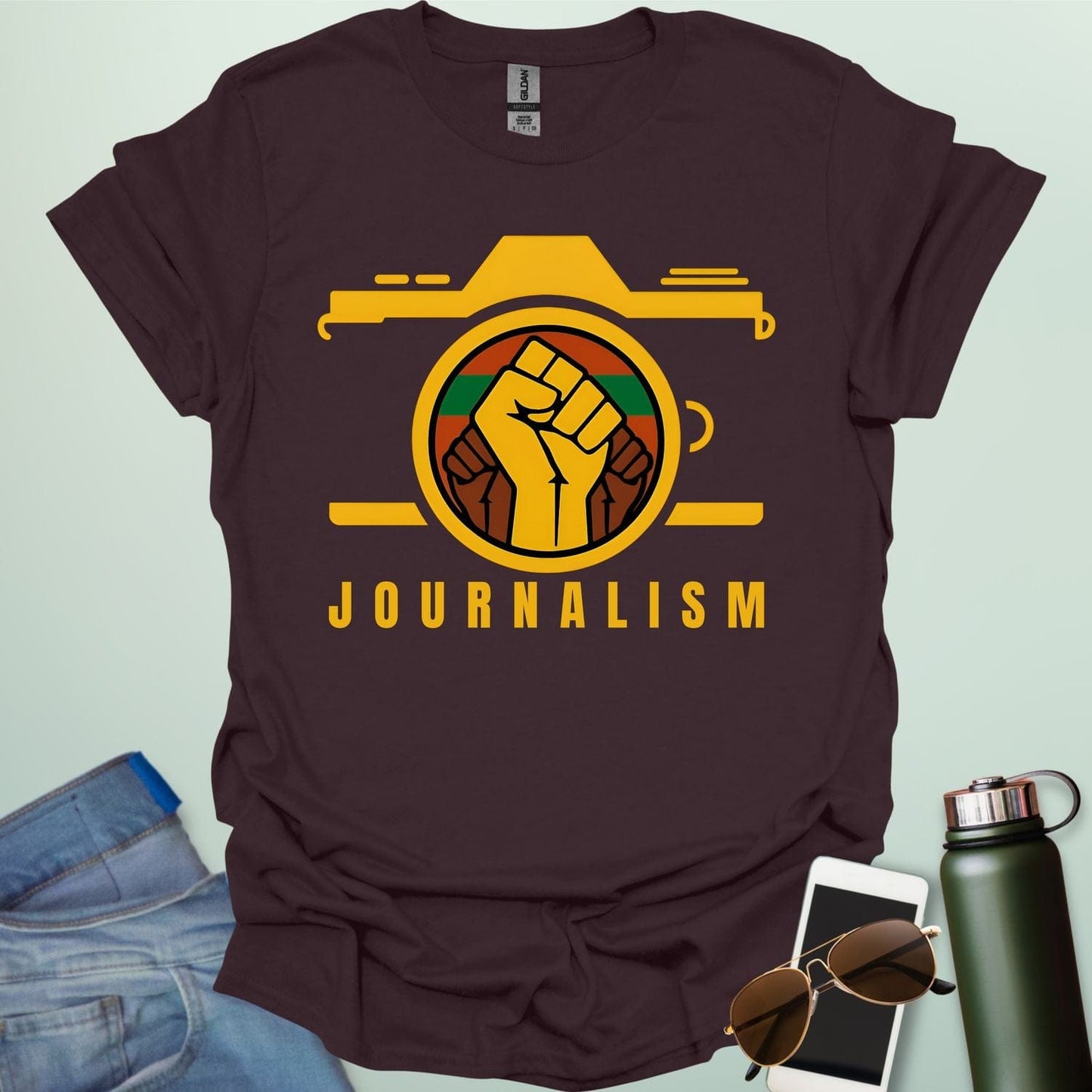 A graphic t-shirt design showing a stylized camera with a powerful raised fist inside its lens. The fist has a green, red, and black striped background. Below the image, the word "JOURNALISM" is written in yellow capital letters.