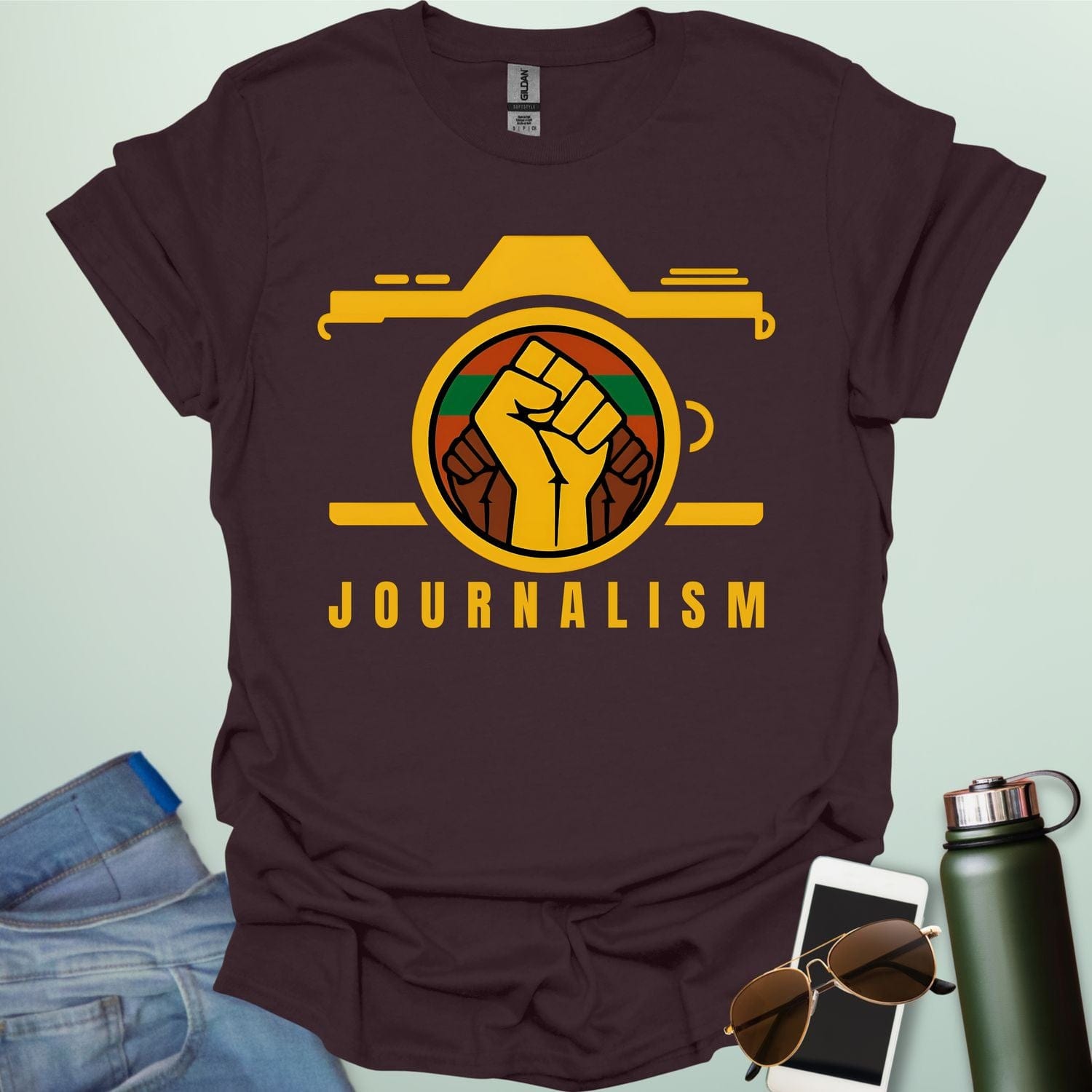 A graphic t-shirt design showing a stylized camera with a powerful raised fist inside its lens. The fist has a green, red, and black striped background. Below the image, the word "JOURNALISM" is written in yellow capital letters.