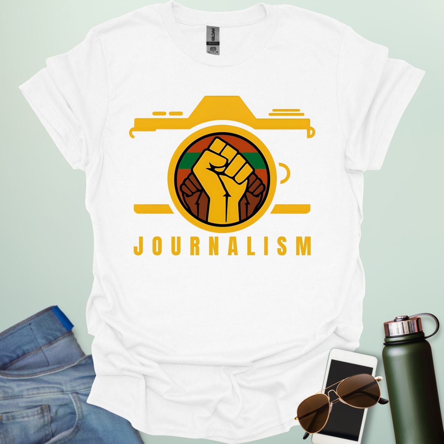 A graphic t-shirt design showing a stylized camera with a powerful raised fist inside its lens. The fist has a green, red, and black striped background. Below the image, the word "JOURNALISM" is written in yellow capital letters.