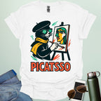 A white t-shirt with a cartoon of a cat painting a Picasso-style portrait on an easel and the text 'PICATSSO' below.
