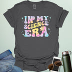 A charcoal grey t-shirt with wavy, tie-dye text that says 'IN MY SCIENCE ERA' with flowers and stars.