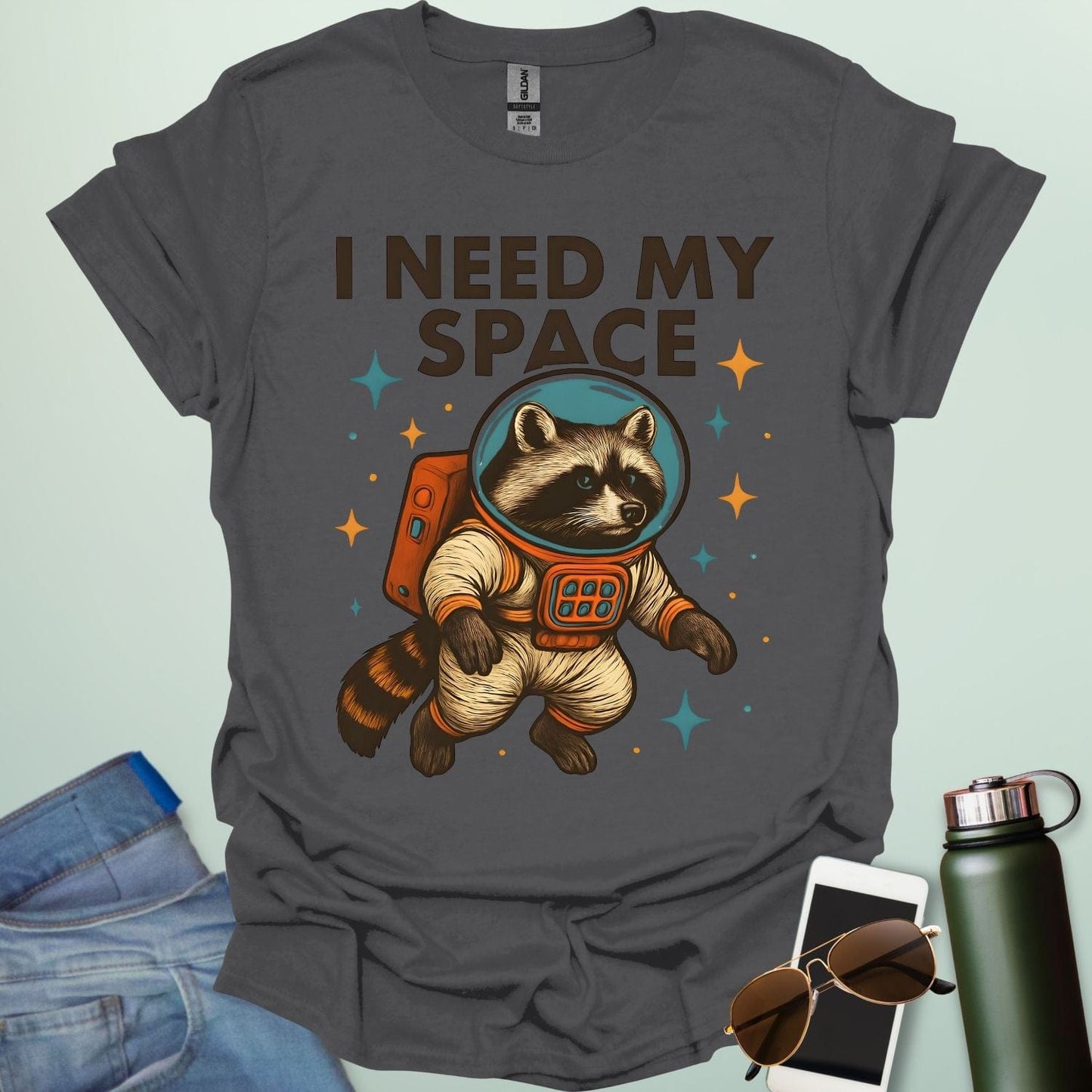 A charcoal grey t-shirt with a cartoon illustration of a raccoon in an astronaut suit and the text 'I NEED MY SPACE.'