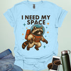 A light blue t-shirt with a cartoon illustration of a raccoon in an astronaut suit and the text 'I NEED MY SPACE.'3. Alt Text