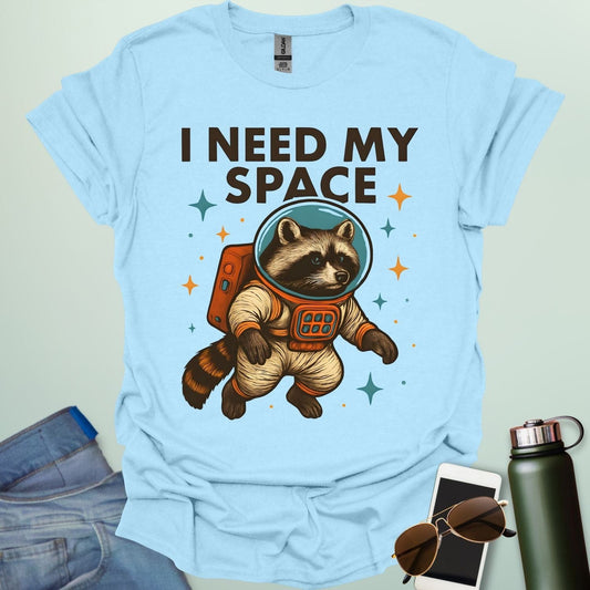 A light blue t-shirt with a cartoon illustration of a raccoon in an astronaut suit and the text 'I NEED MY SPACE.'3. Alt Text